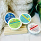 Three shower tabs in white packaging labeled 'Time Out', 'Eucalyptus', and 'Rosemary', placed on a custom wooden soap dish, with a white fluffy towel and a green plant in the background.