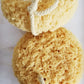 Two large round yellow sponges tied with twine, intended for lathering, displayed on a white surface.