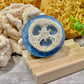 A bar of handcrafted soap with a blue and natural brown color, placed on a wooden dish with a textured background.