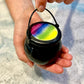 A hand holding a black bath bomb with a colorful, swirling lid resembling a sorting hat.