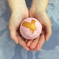 A person holding a gold-dusted bath bomb with a cross-shaped pattern on it, with a blurred background.