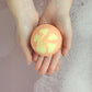 A person holding a orange and pink bath bomb in their hands, with bath bubbles in the background.
