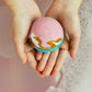 A person holding a colorful bath bomb with layers of pink, blue, and green, topped with a pink and blue powdered sugar coating.