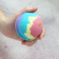 A multicolored bath bomb held in a person's hand, displaying layers of pink, blue, and yellow with a gold unicorn horn charm on top.