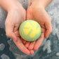 A person holding a green and yellow bath bomb in their hands, with bubbles in the background.