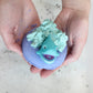 A hand holding a blue bath bomb bomb with a playful shark face design on top, with a backdrop of glitter and loose tea leaves.