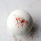 A white bath bomb with a dusting of red spices on its surface, set against a white background.