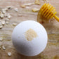A white bath bomb with milk and honey on it, placed on a wooden surface with honey and seeds around, and a honey dipper in the background.