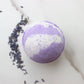A purple and white bath bomb on a white surface with some lavender sprigs scattered around it.