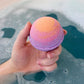 A hand holding a colorful bath bomb with a gradient of orange, yellow, and pink, with a blurred background of bubbles in a bath of water.