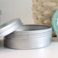 A shampoo bar next to a metal tin, with the lid open and placed next to a woven basket. 