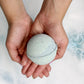 A person holding a bath bomb in their hands, with water bubbles around, indicating the bath bomb is being used in a bath.