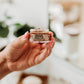 A close-up image of a person's hand holding a small, rectangular container of Chocolate Wasted Sugar Lip Scrub.