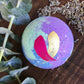 A multicolored bath bomb with purple, green, and gold layers, placed on a wooden surface with dried herbs and plants in the background.